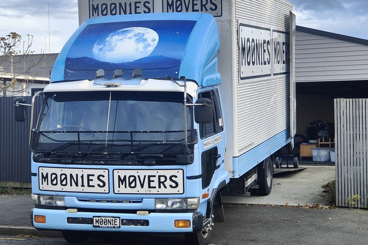 Moonies Movers' light blue removal truck parked on a driveway, MOONIES MOVERS number-plate logo on the front and side of the box