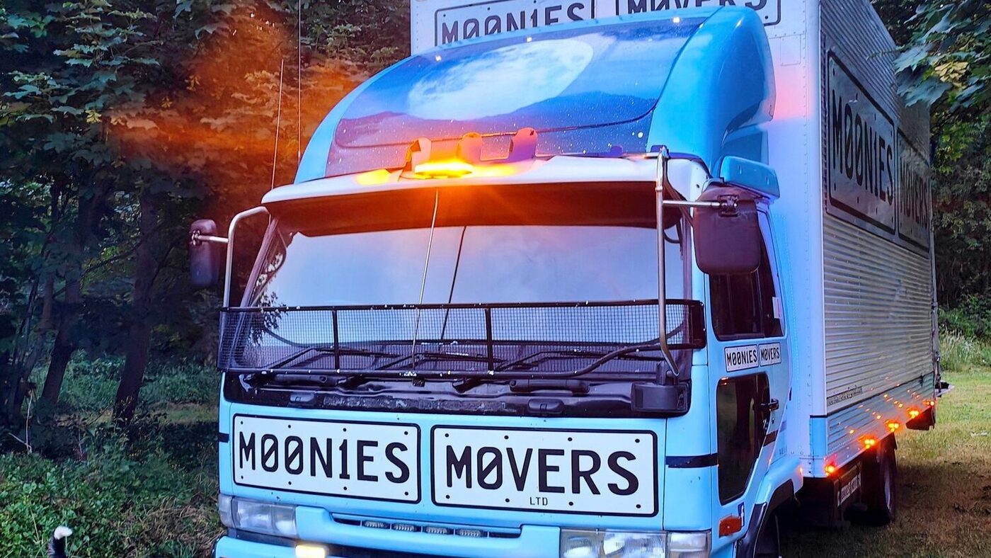 The Moonies Movers truck at dusk with its amber beacon on and the MOONIES MOVERS logo across the front number plates