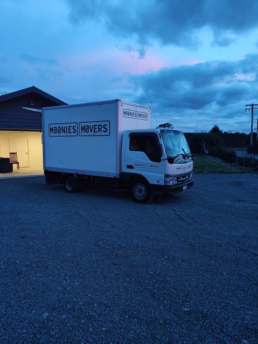 Moonies Movers' white truck parked on a gravel drive at blue hour with the house lights on behind it