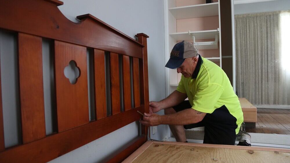 A removalist in a high-visibility shirt taking a solid timber bed frame apart in a bedroom, ready to carry it out