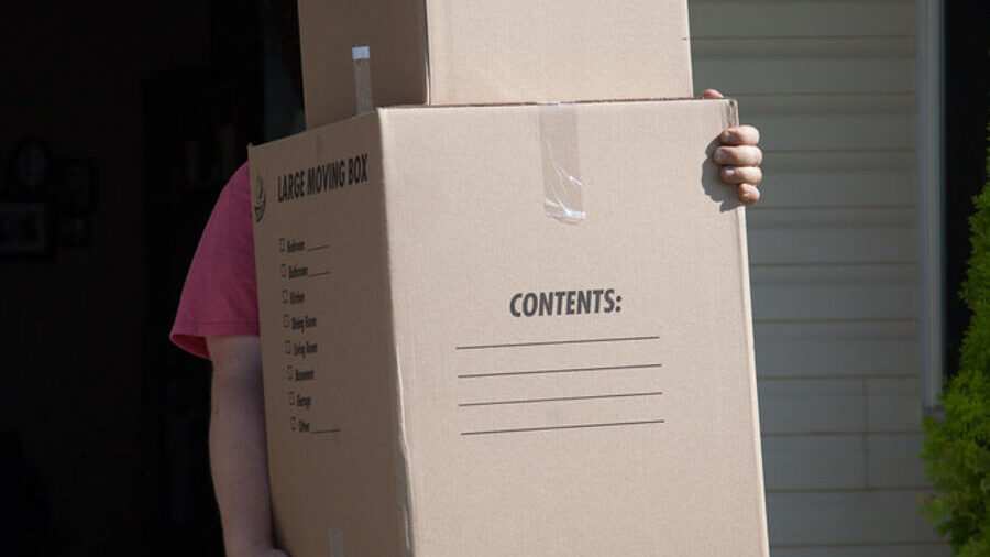 A person carrying two stacked moving boxes out through the front door of a weatherboard house