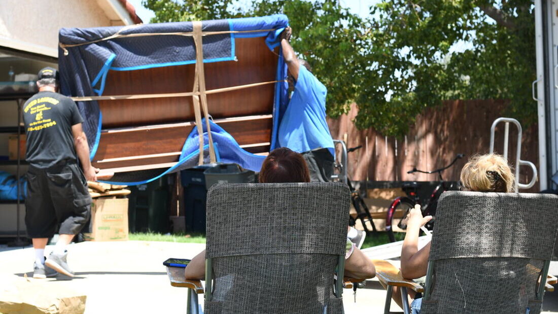 Two people carrying a mattress wrapped in a blue moving blanket past neighbours watching from garden chairs