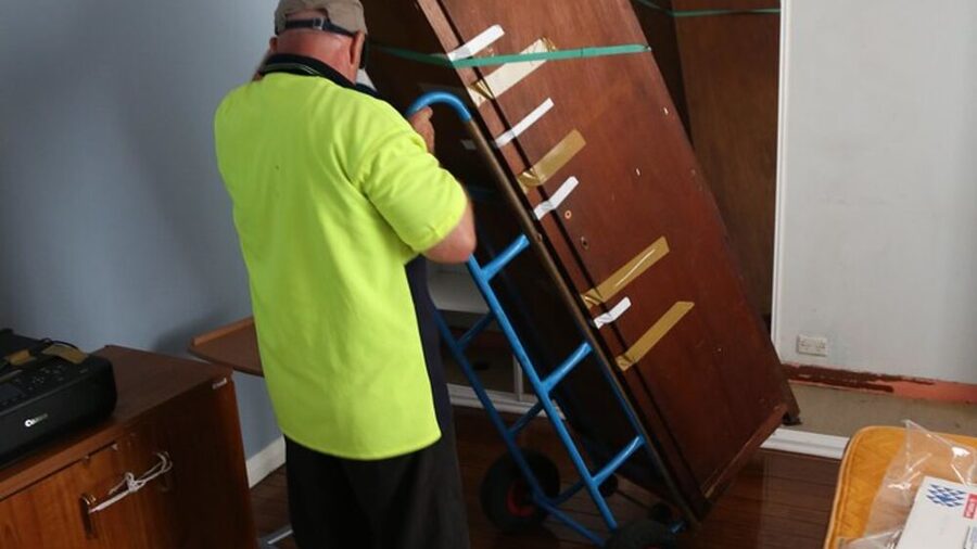 A removalist in a high-visibility shirt wheeling a large timber wardrobe on a hand truck through a hallway
