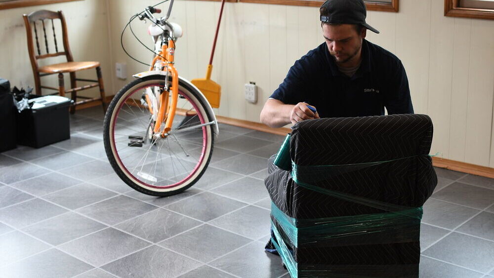 A mover shrink-wrapping a dark armchair in green stretch film in a sunroom before it goes on the truck